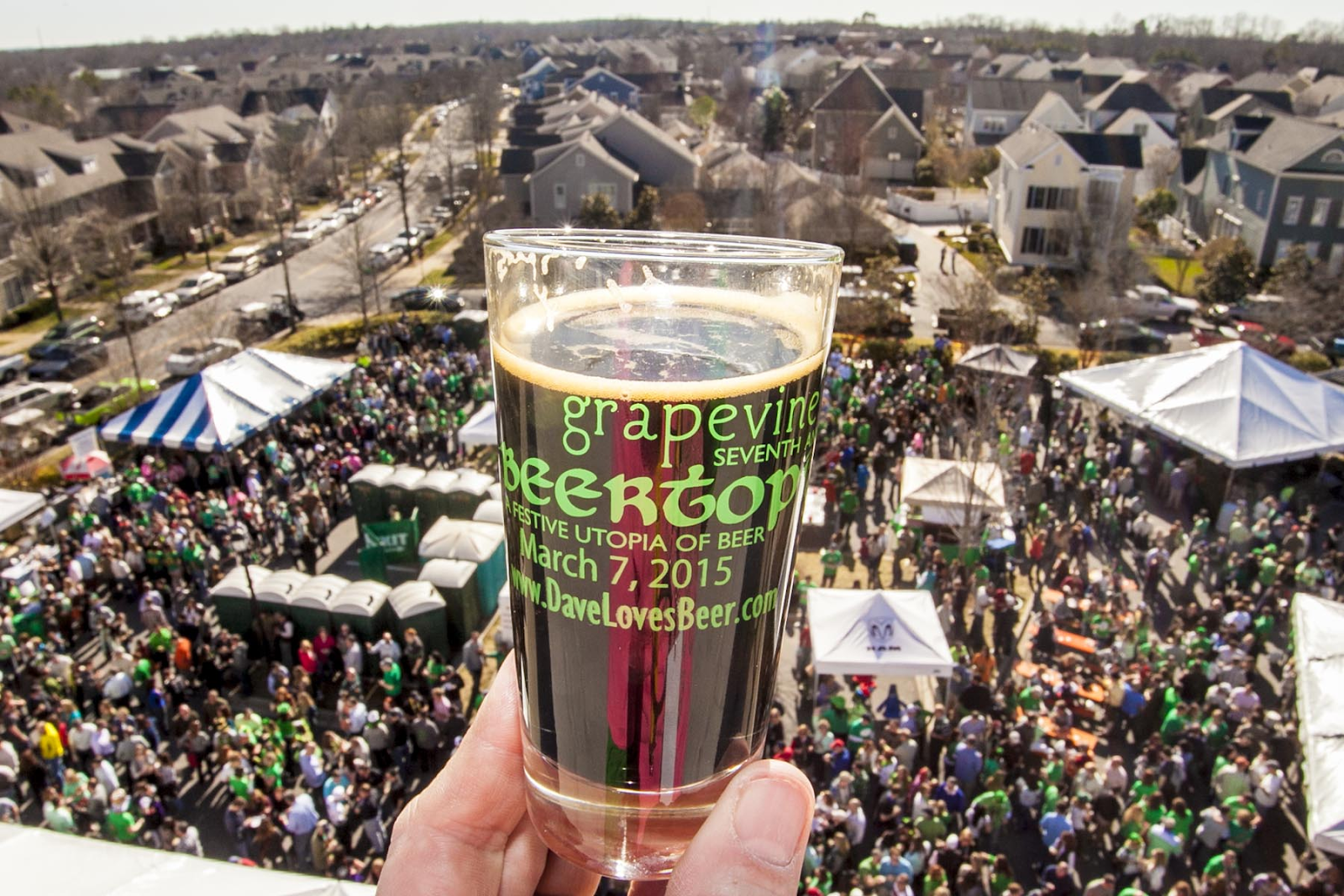 A hand holding a glass of beer over a view of people having fun at Grapevine’s Beertopia
